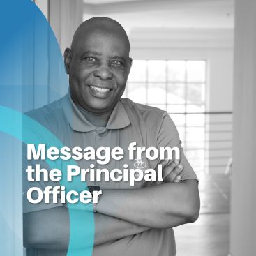 Portrait of the Principal Officer smiling with arms crossed, standing in a bright office space.
